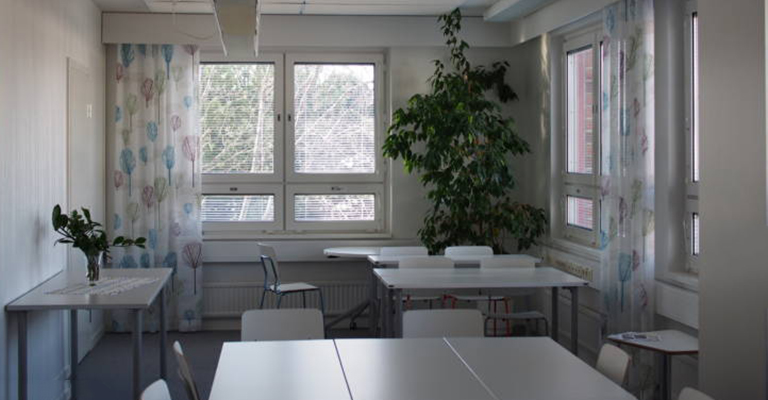Kuva Vääksyn toimistotiloista. Kuvassa valkoisia pöytiä ja tuoleja sekä iso sisäkasvi nurkassa ja ikkuna, jossa vaalea verho.