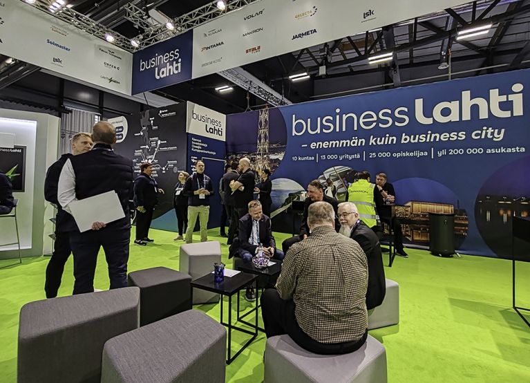 Lahden seudun yhteisosasto Alihankinta 2021 Team Lahti Business Region messuseinän edessä. Seinässä teksti: huippusuorituksiin Lahden seudulla.