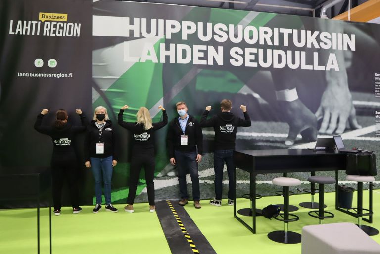 Team Lahti Business Region messuseinän edessä. Seinässä teksti: huippusuorituksiin Lahden seudulla.