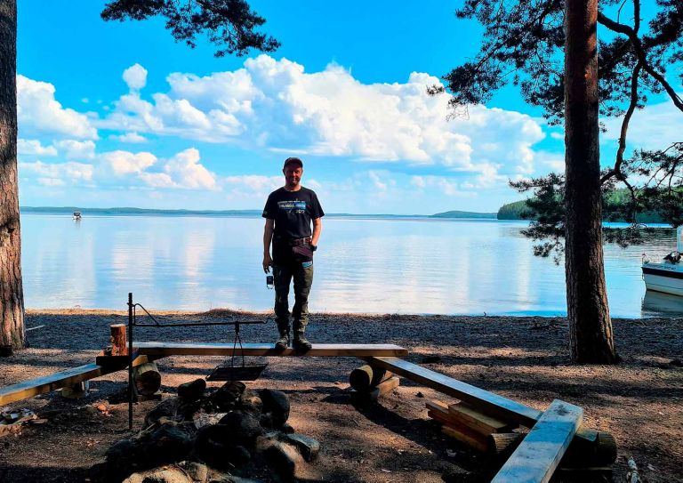 Kuva: Päijänteen luonnonpuisto, Kelventeen saari, VisitLahti.fi A man standing on a bench by a campfire. A lake in the background.