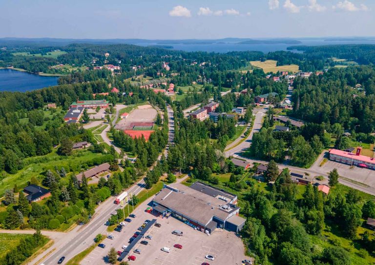 Aurinkoinen ilmakuva. Paljon vihreää metsää, teiden varrella rakennuksia. Etualalla iso rakennus ja parkkipaikka.