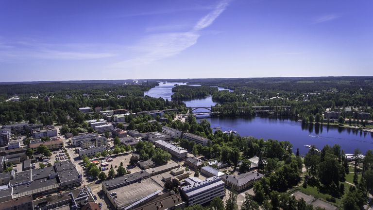 Kuvat: VisitLahti.fi A deep blue sky. The center of Heinola, with buildings and a market square. In the background, a river and a bridge.